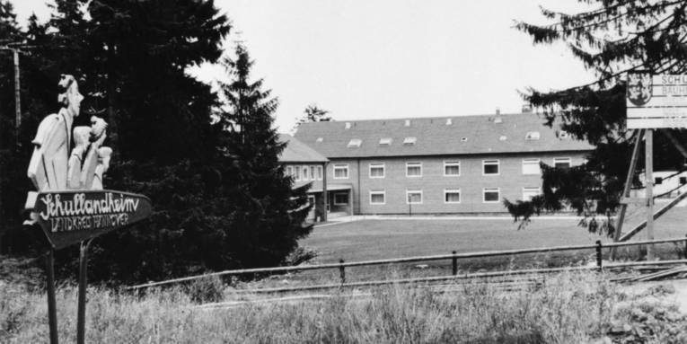 Schwarz-Weiß-Fotografie: Wegweiser mit geschnitzten Köpfen und dem Text "Schullandheim Torfhaus", im Hintergrund ist ein Gebäudekomplex zu sehen.