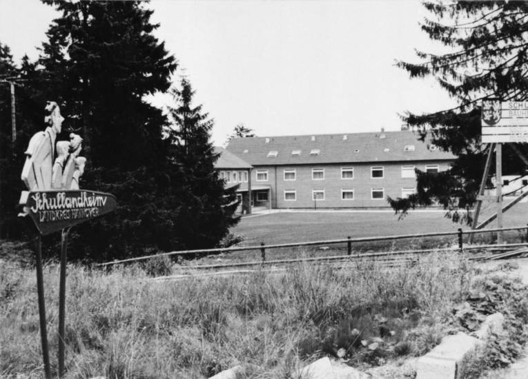 Schwarz-Weiß-Fotografie: Wegweiser mit geschnitzten Köpfen und dem Text "Schullandheim Torfhaus", im Hintergrund ist ein Gebäudekomplex zu sehen.