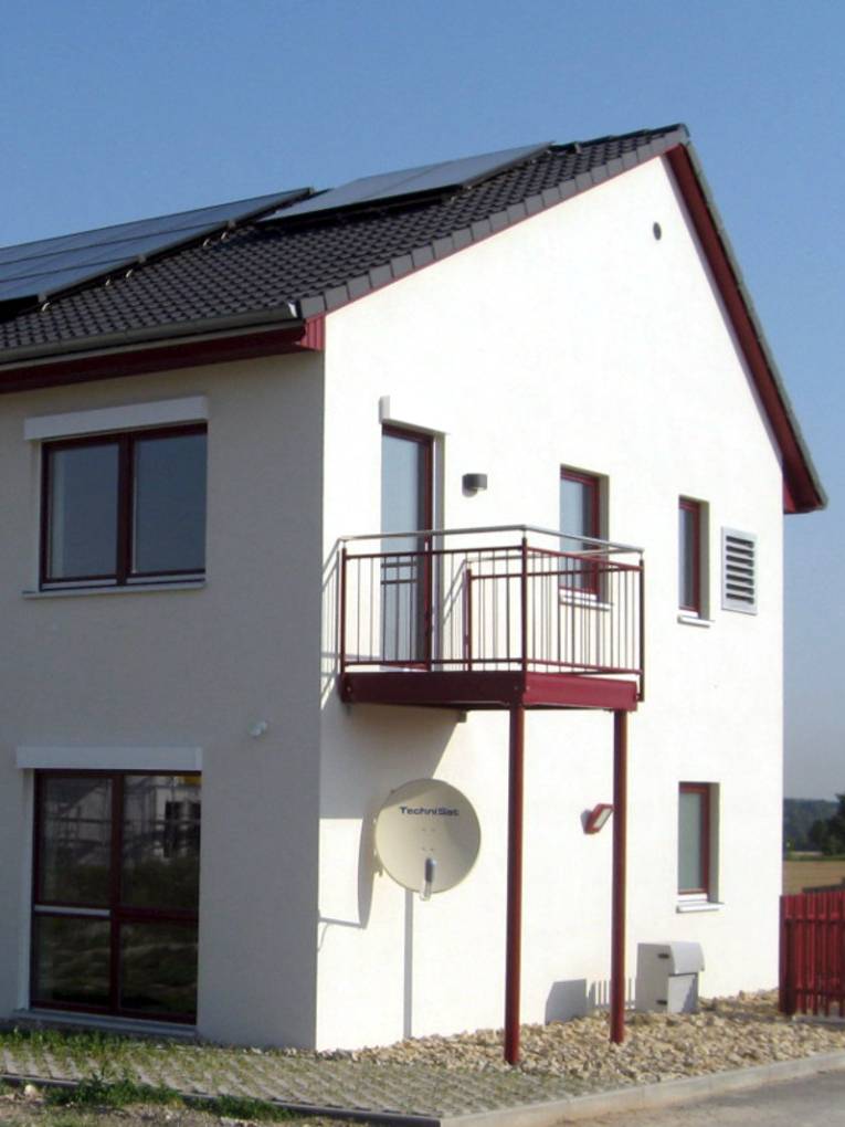 Einfamilienhaus mit weißer Fassade, großen Fenstern und einem Balkon. Auf dem Dach ist eine Solaranlage, Photovoltaikzellen erzeugen Strom, Solarthermie sorgt für Wärme.