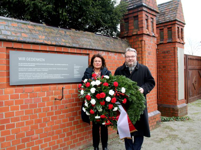 Zwei Personen bei einer Kranzniederlegung vor einer Gedenkwand.