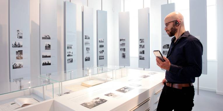 Ein Mann steht in einer hellen Bilder-Ausstellung, trägt ein Gerät in den Händen und einen Knopf im Ohr.