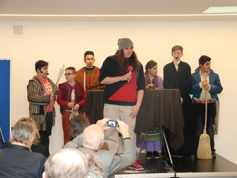 Eine Schülerin und sechs Schüler auf einer kleinen Bühne. Ein Junge steht vorne mit einem Handmikrofon - alle anderen tragen ein Kostüm.