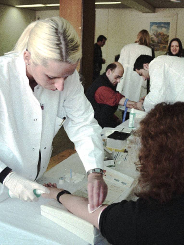 Eine Frau hat ihren linken Arm ausgestreckt auf einem Polster abgelegt, eine Frau in einem weißen Kittel desinfiziert die Armbeuge. Im Hintergrund spielt sich eine ähnliche Szene zwischen zwei Männern ab, weitere Menschen warten im Hintergrund.