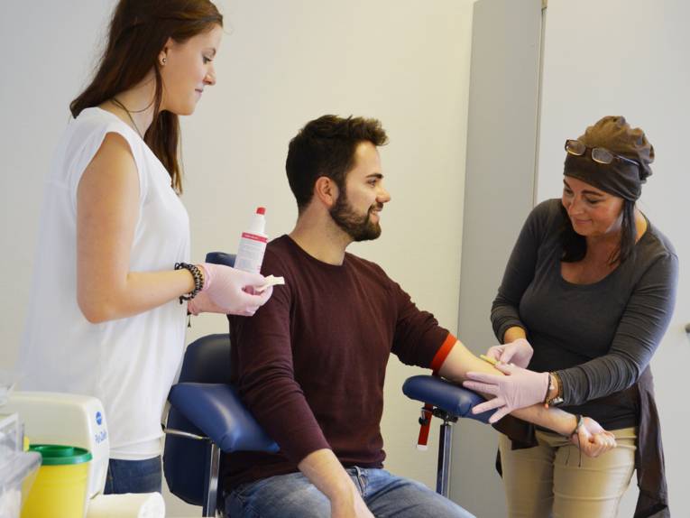 Drei Personen, eine Frau mit hält einen Behälter mit Desinfektionsmittel in der Hand, ein Mann sitzt auf einem Stuhl und lässt sich aus dem linken Arm Blut abnehmen, eine Frau nimmt dem Mann Blut ab