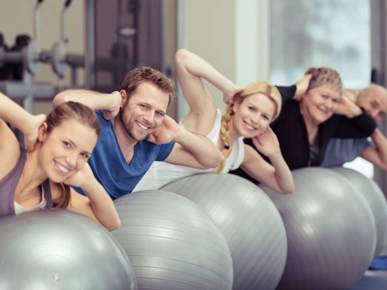 Eine Gruppe von Personen unterschiedlicher Altersgruppen trainiert in einem Fitnessraum mit großen Gymnastikball