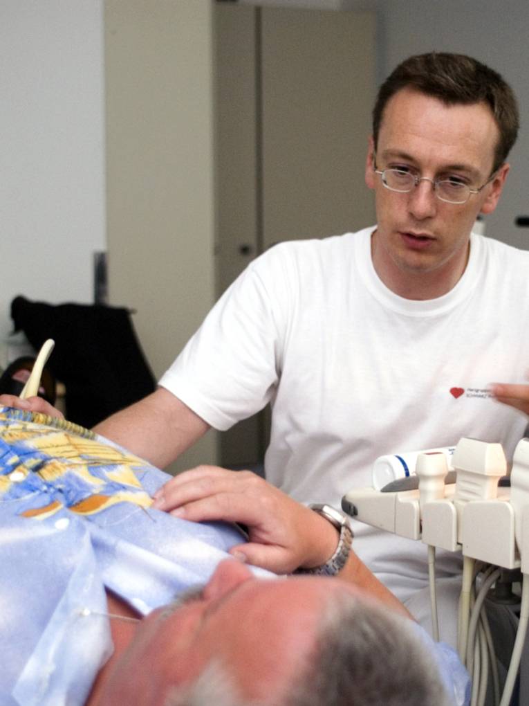 Ein Mann fährt mit einem Ultraschallkopf über den Unterleib eines liegenden, älteren Mannes.
