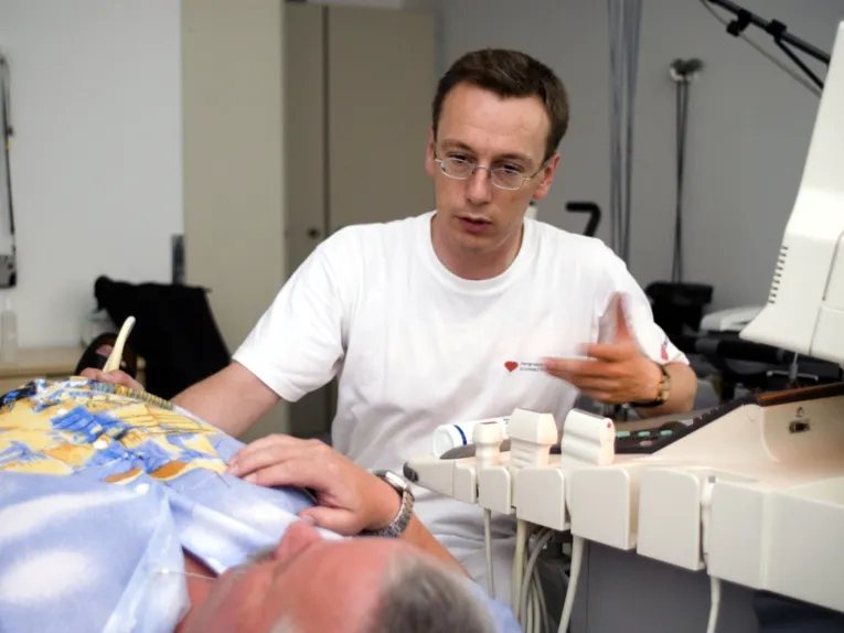 Ein Mann fährt mit einem Ultraschallkopf über den Unterleib eines liegenden, älteren Mannes.