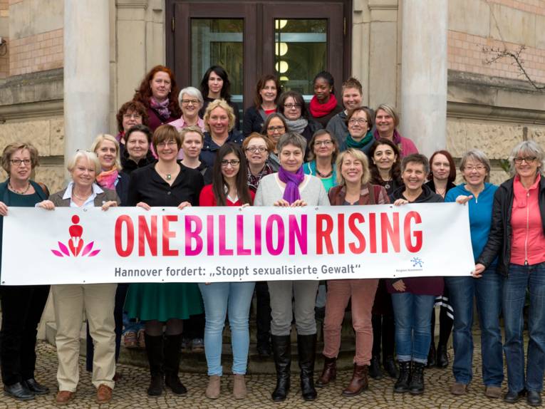 28 Frauen stehen auf einer Treppe und schauen in die Kamera, die Frauen in der vordersten Reihe halten ein Banner, darauf steht: "ONE BILLION RISING. Hannover fordert: 'Stoppt sexualisierte Gewal'"