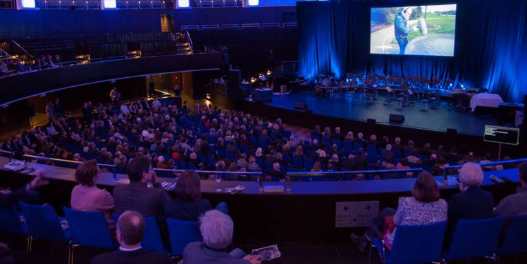 Publikum in einem großen Veranstaltungssaal mit mehreren Reihen und Rängen. Die Gäste schauen in Richtung Bühne, auf der über eine Leinwand ein Film abgespielt wird.