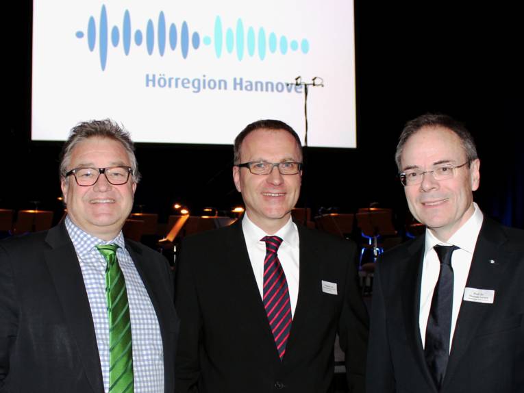 Drei Männer stehen nebeneinander, im Hintergrund ist eine Leinwand, auf die das Logo der Hörregion Hannover projeziert wird,