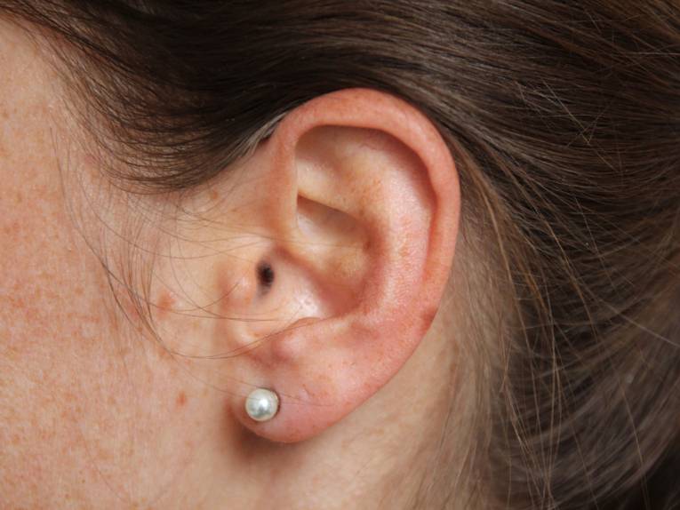 Das Ohr einer dunkelhaarigen Frau mit einem Perlenohrstecker.