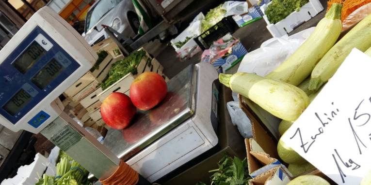 Ein Obst- und Gemüsestand auf einem Wochenmarkt. Auf der Waage liegen zwei rote Äpfel und Zuchini steht zum Verkauf.