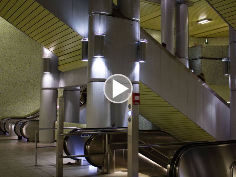 Unterirdische Stadtbahnstation mit mehren Rolltreppen. In der Mitte des Bildes ist ein Abspielsymbol abgebildet. 