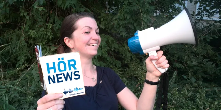Eine Dame hält ein Heft mit der Aufschrift "Hörnews" in der Hand und ruft etwas in Richtung eines Megafons