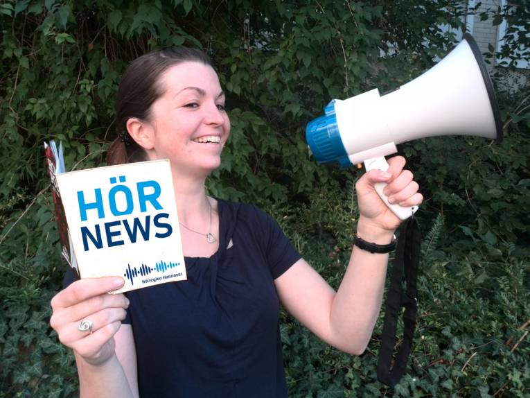 Eine Dame hält ein Heft mit der Aufschrift "Hörnews" in der Hand und ruft etwas in Richtung eines Megafons
