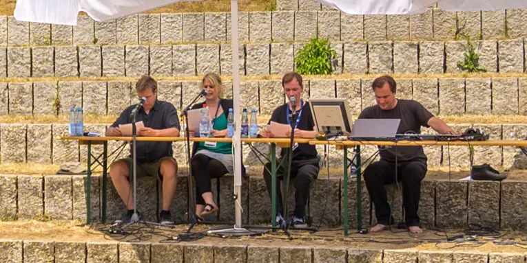 Vier Personen in schwarzen T-Shirts sitzen im Freien und sprechen in Mikrofone vor ihnen.