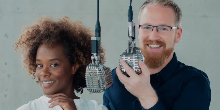 Eine Frau mit Locken und ein Mann mit Bart und zwei Mikrofone im Vordergrund.