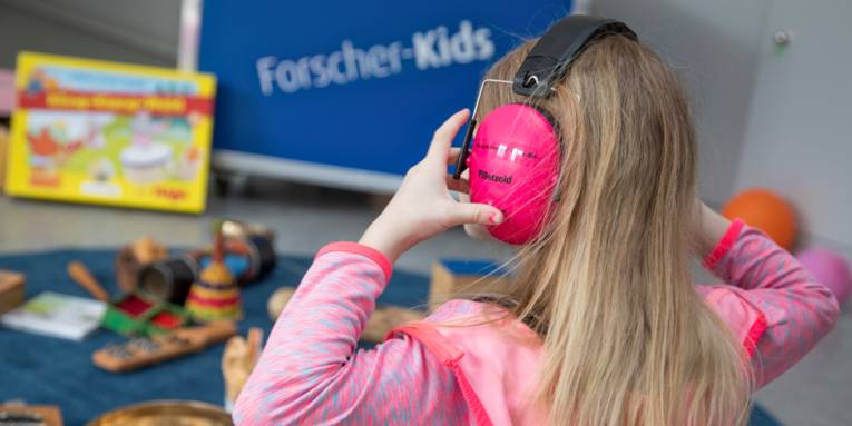 Ein Mädchen trägt Kopfhörer, im Hintergrund ist die Entdeckerkiste der Forscherkids zum Thema Klänge und Geräusche zu sehen, Instrumente und andere klingende Gegenstände oder Material für Hör-Experimente liegt daneben.