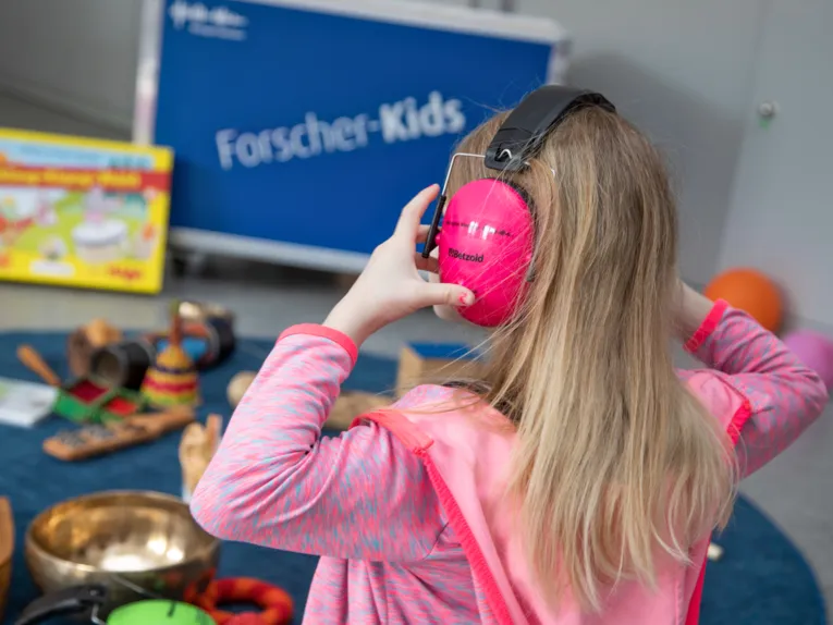 Ein Mädchen trägt Kopfhörer, im Hintergrund ist die Entdeckerkiste der Forscherkids zum Thema Klänge und Geräusche zu sehen, Instrumente und andere klingende Gegenstände oder Material für Hör-Experimente liegt daneben.
