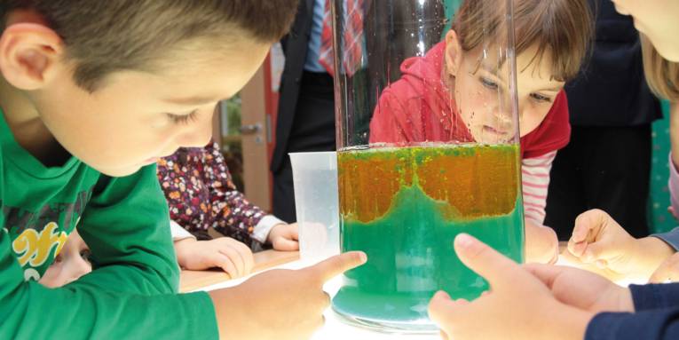 Kinder, die schauen auf einen mit gelber und grüner Flüssigkeit gefülltem durchsichtigen Behälter