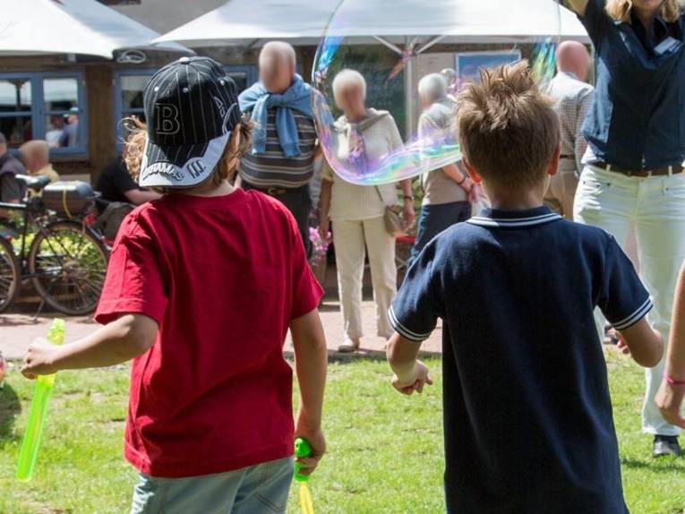 Zwei Kinder stehen auf einer Rasenfläche und beobachten eine Frau, die Seifenblase macht.