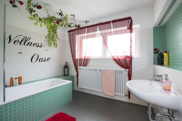 Badezimmer mit t&uuml;rkis gefliester Badewanne und dunklen Fu&szlig;bodenfliesen. An einer Wand steht "Wellness Oase" geschrieben und Rosen sind aufgemalt. In zwei Ecken sind Rosen als Dekoration aufgestellt. Auf dem Waschbecken steht ein Seifenspender, auf der Ablage &uuml;ber dem Waschbecken ein Reinigungsmittel und bei der Badewanne befinden sich Schampoo, Duschgel und Sp&uuml;lung. Auf dem Boden und auf der Heizung liegt jeweils ein Handtuch.