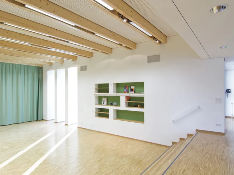 Ein saalähnlicher Raum mit hellem Holzfußboden und wenigen Stufen. In einer Wand sind sechs Regale, deren Rückwände grün gestrichen sind.