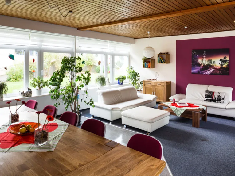Ein Raum ausgestattet mit zwei weißen Sofas, einem Holzcouchtisch, einem kleinen Kommodenschrank, ein großen Esstisch mit rot gepolsterten Stühlen an der einen Tischseite, zwei kleinen Bücherregalem und einem großen Bild an der Wand sowie mehreren Grünpflanzen.