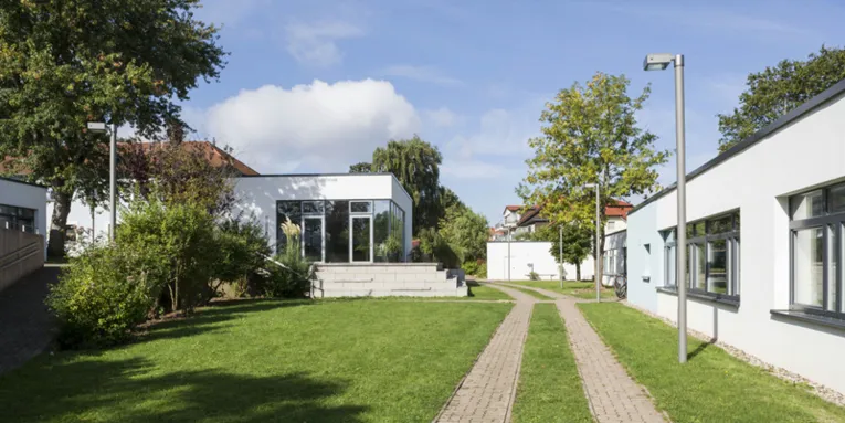 Mehrere kleine, weiß verputzte Häuser mit Flachdach und dazwischen grüne Rasenfläche. Auf dem Gelände befinden sich außerdem mehrere Bäume und Straßenlaternen.