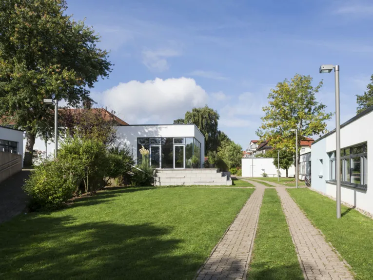 Mehrere kleine, weiß verputzte Häuser mit Flachdach und dazwischen grüne Rasenfläche. Auf dem Gelände befinden sich außerdem mehrere Bäume und Straßenlaternen.