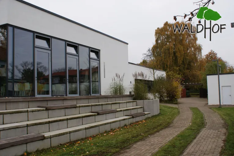 Ein wei&szlig;es Geb&auml;ude mit Flachdach. Davor sind Treppenstufen, die so breit sind wie das Geb&auml;ude an sich und als Sitzb&auml;nke (Trib&uuml;ne) verwendet werden k&ouml;nnen.
