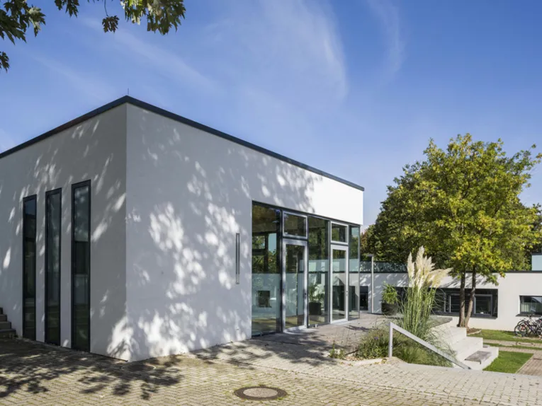 Ein wei&szlig;es Geb&auml;ude mit Flachdach. Davor sind Treppenstufen und ein Gel&auml;nder. Dahinter ist ein weiteres Haus mit Flachdach zu sehen, vor dem ein gro&szlig;er Baum steht und Fahrr&auml;der abgestellt sind.