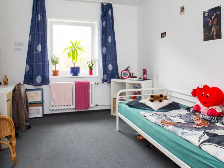 Ein Jugendzimmer mit einem Bett, auf dem neben Bettzeug mehrere Kuscheltiere und ein Herzkissen liegen. An der Wand sind ein Poster und zwei Fotos angebracht. Auf der Heizung liegen zwei Handt&uuml;cher und in der Fensterbank stehen drei Pflanzen. Au&szlig;erdem stehen im Zimmer ein Schreibtisch, ein Schreibtischstuhl, ein weiterer Stuhl und ein kleines Stehregal.
