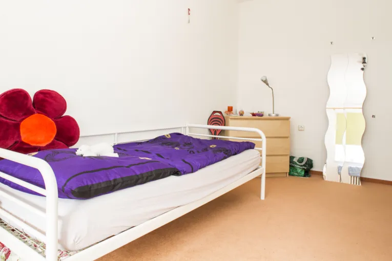 Ein Jugendzimmer mit einem Bett, auf dem eine Bettdecke, ein Kopfkissen und ein Blumenkissen liegen. An die Wand ist ein Spiegel gelehnt und daneben steht eine Kommode.