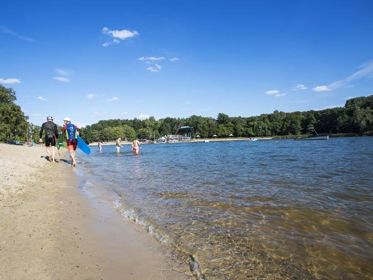 Zwei Surfer gehen einen Strand entlang, in der Ferne baden weitere Personen im Blauen See Garbsen. Im Hintergrund sind Buden, Zelte und Sonnenschirme, im Wasser sind Hindernisse der Wasserskianlage.