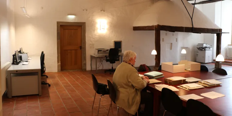 Ein Mann sitzt an einem großen Tisch, auf dem viele Blätter bzw. Dokumente verteilt liegen.