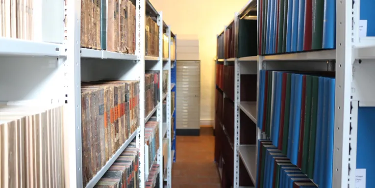 Zwei Regalreihen mit Büchern und Ordner und dazwischen ein Durchgang.