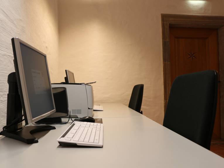 Zwei nebeinander stehende Schreibtische mit Schreibtischstuhl, auf denen sich jeweils ein Monitor und eine Tastatur befinden. Zwischen den beiden Bildschirmen ist ein drucker platziert.