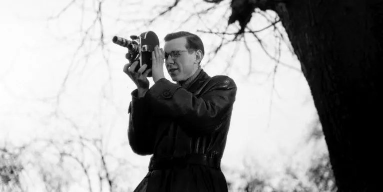 Ein Mann im Ledermantel und mit Brille steht draußen neben einem Baum und fotografiert mit einer Kamera.