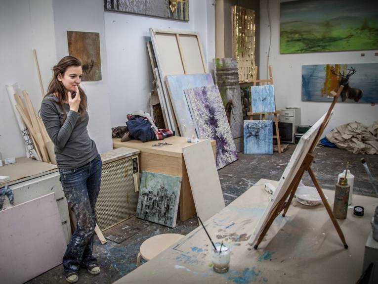 Eine junge Künstlerin in ihrem Atelier. Sie schaut auf eines ihrer Werke auf einer Staffelei. Überall stehen Leinwände in verschiedenen Größen und auch an den Wänden hängen gemalte Bilder.