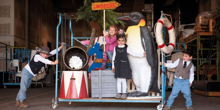 Zwei kleine Jungs rechts und links von einem Rollwagen. Auf dem Rollwagen befinden sich zwei kleine Mädchen und Teile einer Bühnenkulisse (u. a. ein riesiger Pinguin und ein Schaukelpferd).