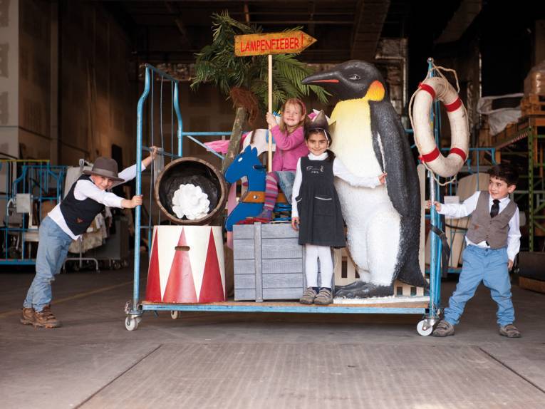Zwei kleine Jungs rechts und links von einem Rollwagen. Auf dem Rollwagen befinden sich zwei kleine Mädchen und Teile einer Bühnenkulisse (u. a. ein riesiger Pinguin und ein Schaukelpferd).