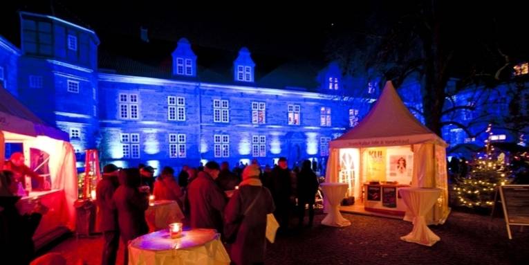 Scheinwerfer tauchen die Fassades von Schloss Landestrost in Neustadt am Rübenberge in ein blaues Licht. Vor dem Schloss sind die Buden eines Weihnachtsmarktes aufgebaut. Menschen wärmen sich an heißen Getränken, am rechten Bildrand ist ein kleiner Tannenbaum mit einer Lichterkette geschmückt.