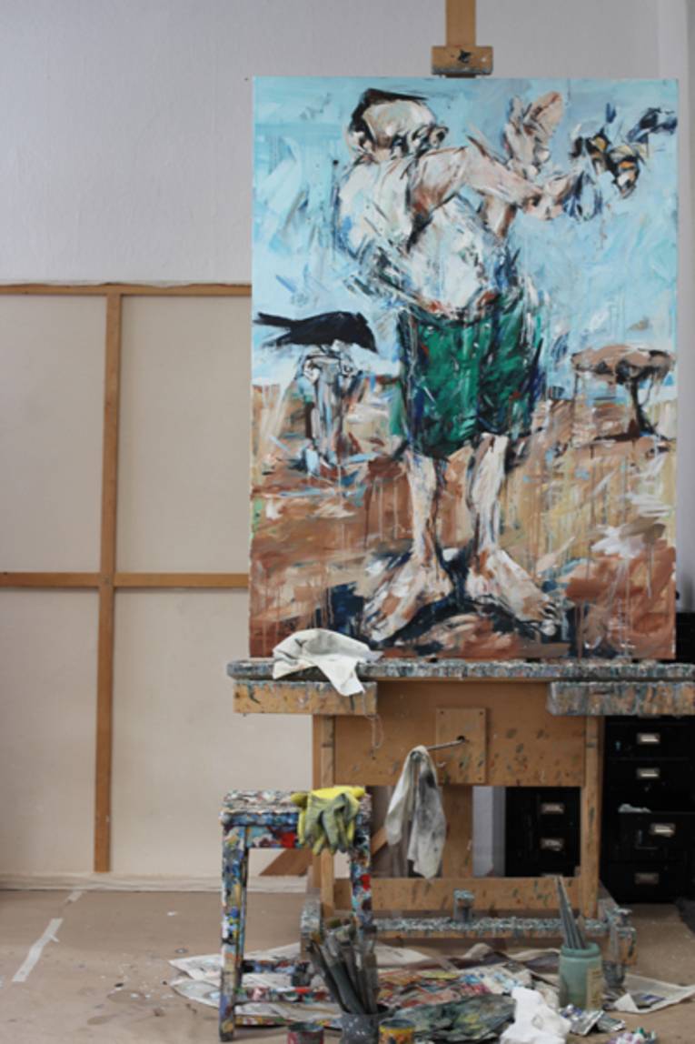 Ein Bild mit einem Mann drauf steht auf einer Staffelei. Davor steht ein. mit Farben beschmierter Hocker und ein Glas mit Pinsel. Auf dem Boden ist Zeitungspapier audsgebreitet.