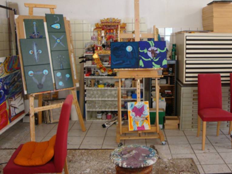 Ein Atelier mit weiß gefliestem Boden. In dem Raum stehen zwei Staffeleien, mehrere Bilder, zwei rote Stühle, ein Hocker, kleine Schränke und ein Regalschrank mit Farben, Pinseln und Malsachen.