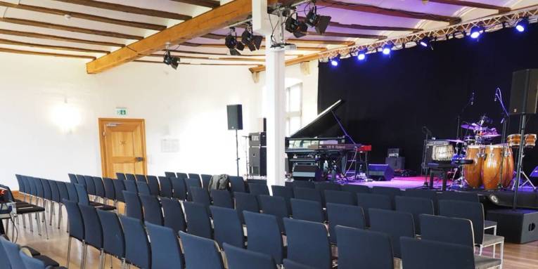 Ein großer Saal in einem Schloss, der als Veranstaltungsraum genutzt wird. Rechts ist eine erhöhte Bühne, auf der Musikinstrumente stehen. Vor der Bühne sind mehrere Stuhlreihen aufgebaut.