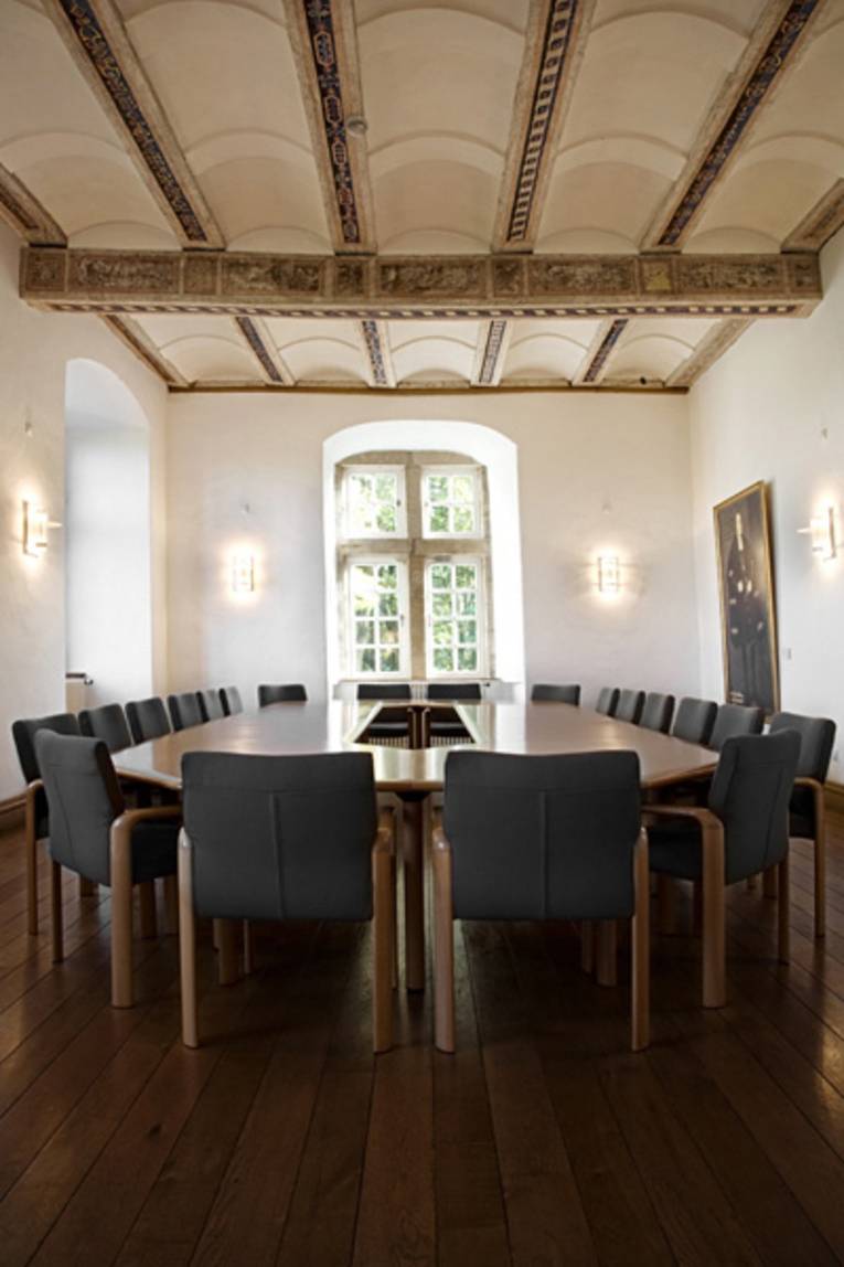 Ein lichtdurchfluteter Saal in einem Schloss mit einem großen Besprechungstisch, andem zwanzig Stühle stehen.