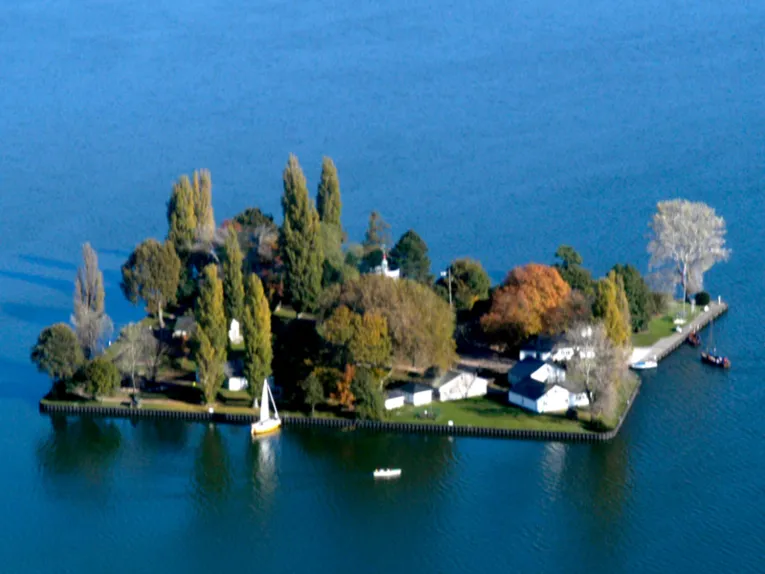 Luftaufnahme: Nahezu quadratische, flache Insel mit B&auml;umen und Geb&auml;uden. Am Ufer ankern zwei Segelbote.