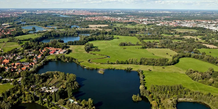Luftaufnahme: Mehrere kleine Gewässer sind durch Gräben miteinander verbunden. Die Gewässer werden unterbrochen von Grünflcäehn, Bäumen und kleinen Siedlungen. Im Hintergrund ist die Stadt Hannover mit Maschsee und Fernsehturm zu erkennen.