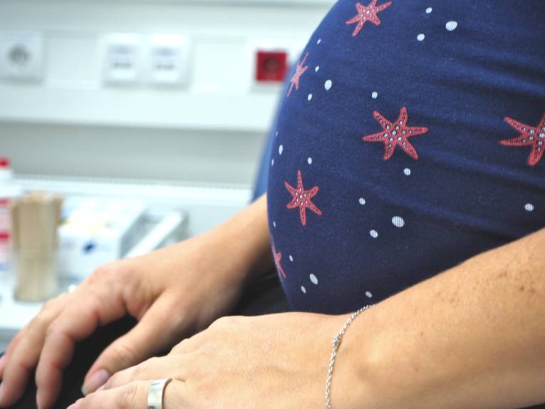 Eine Schwangere sitzt im Behandlungszimmer eines Arztes. Im Bildausschnitt sind ihr Bauch und die Hände, die sie sich in den Schoß legt.
